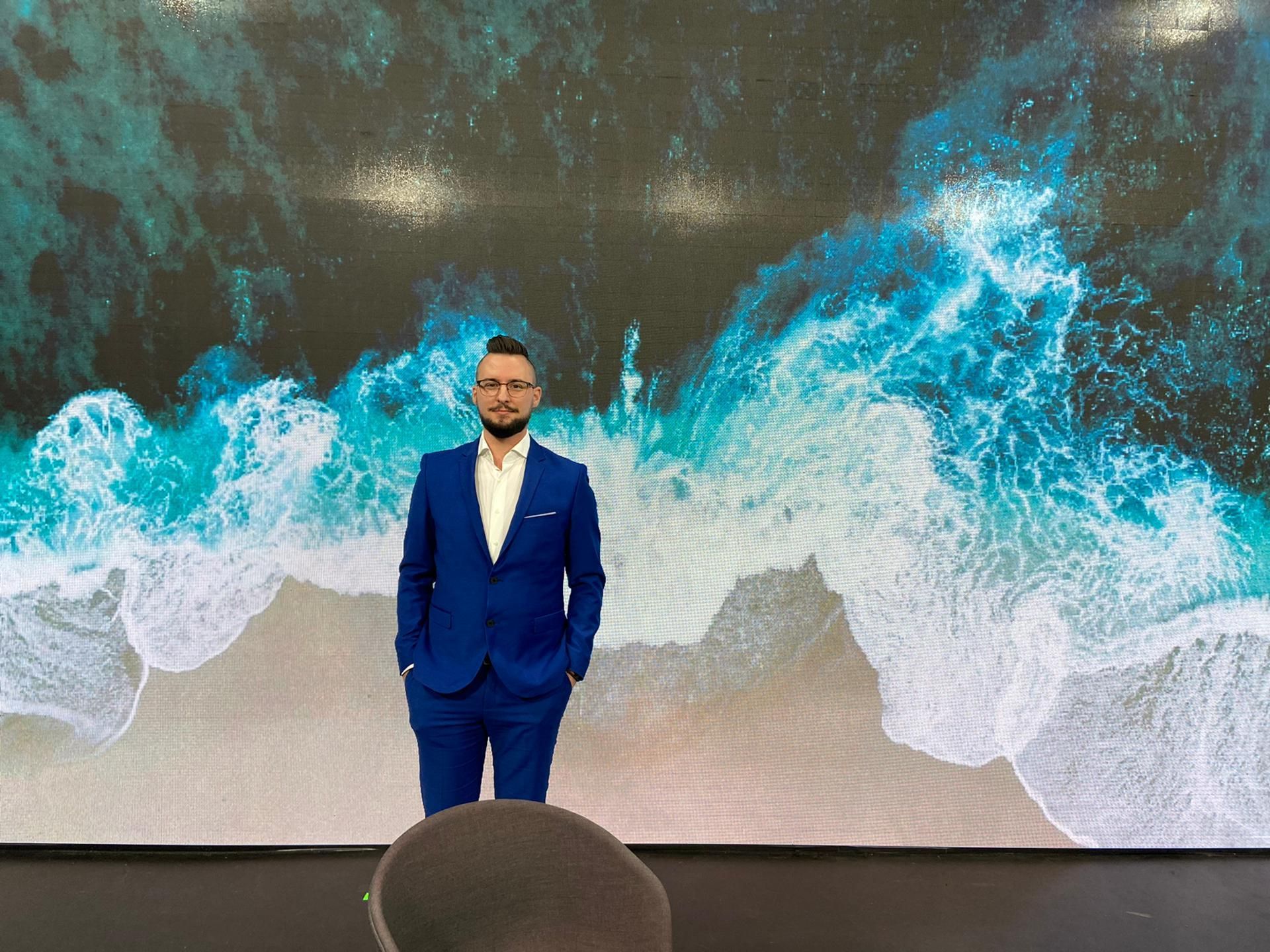 Man in blue suit stands on stage with ocean wave background.