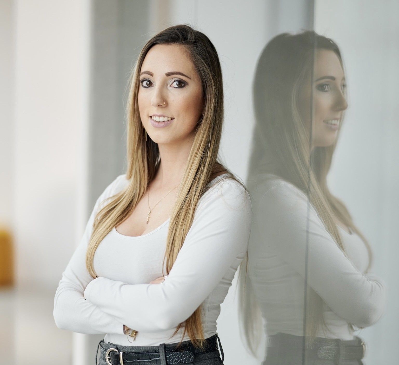 Woman with arms crossed, smiling, leaning against a glass wall. She has long brown hair, wearing a white top and dark pants.