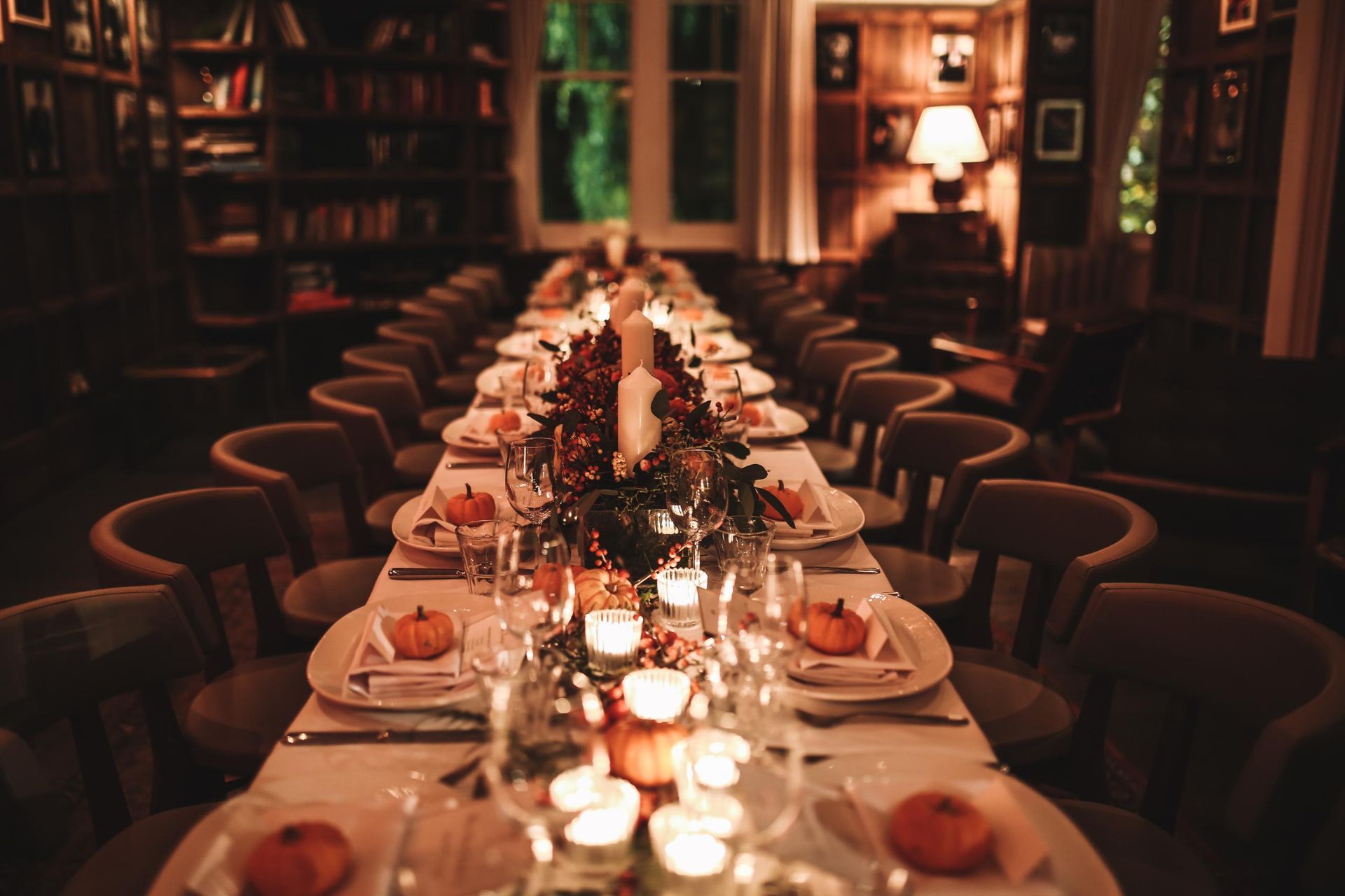 Long table set for a formal dinner with fall decorations, candles, and pumpkins. Dark, cozy room.
