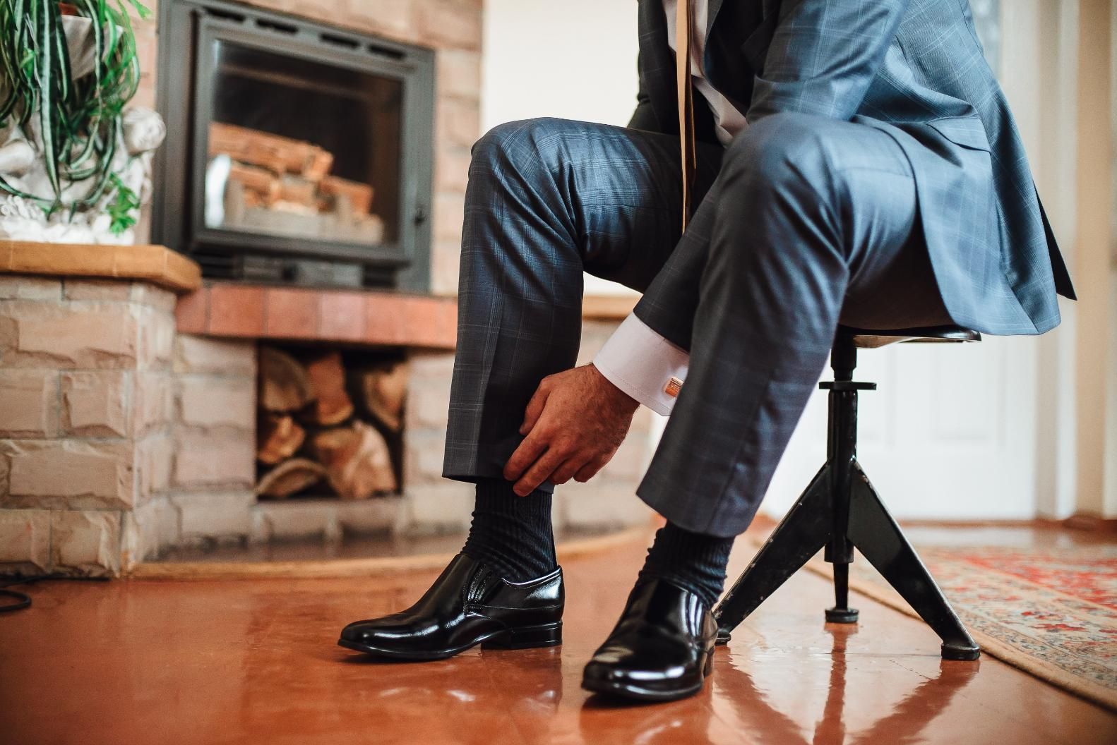 Man in suit adjusting sock, sitting near a fireplace.