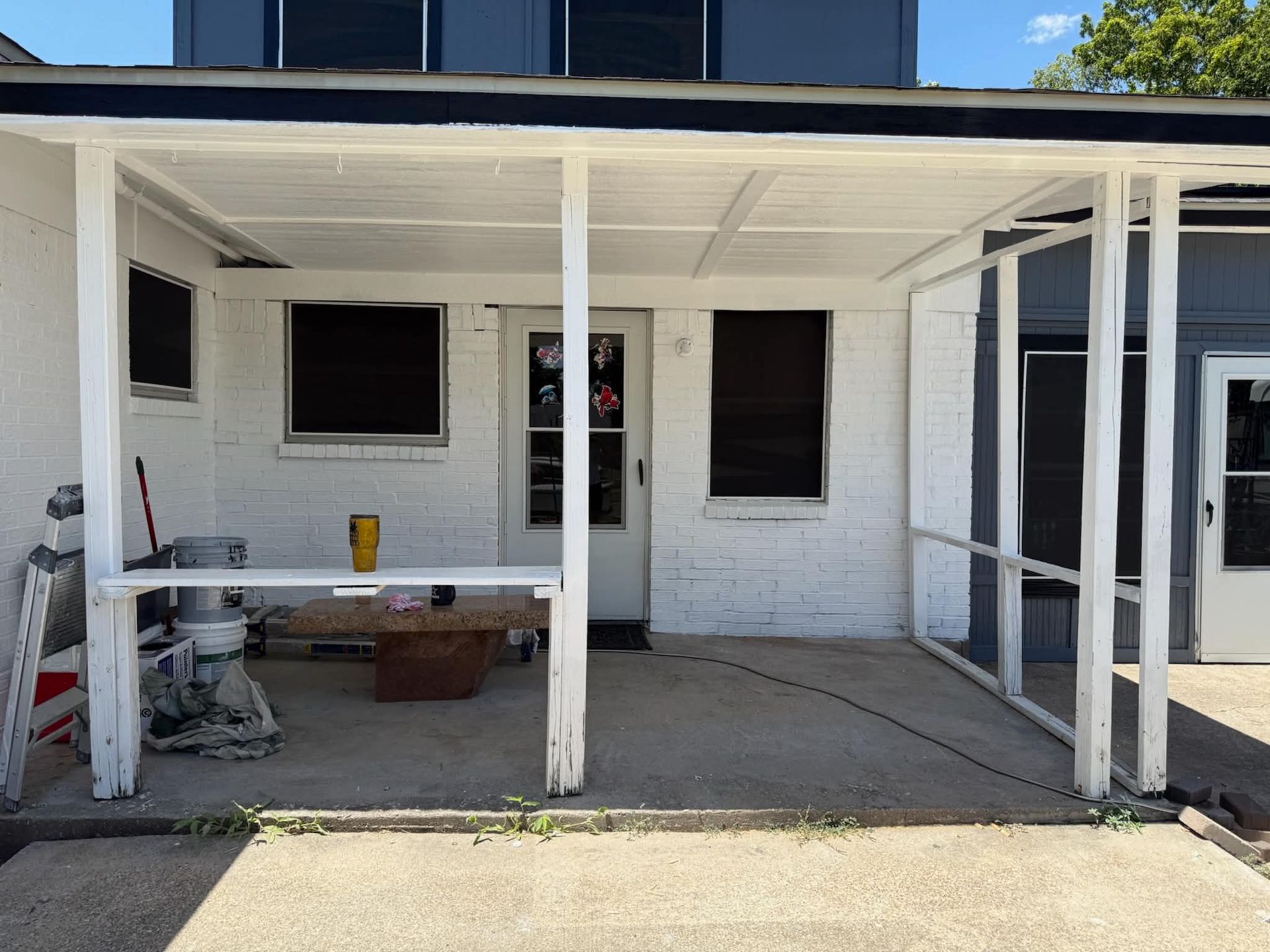 Covered front porch with white posts, three windows, a screen door, and a small work table with tools.