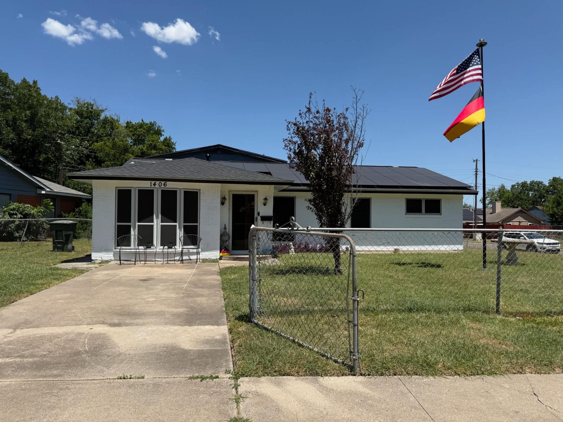 Single-story white house with a driveway, chain-link fence, and two flags in the front yard