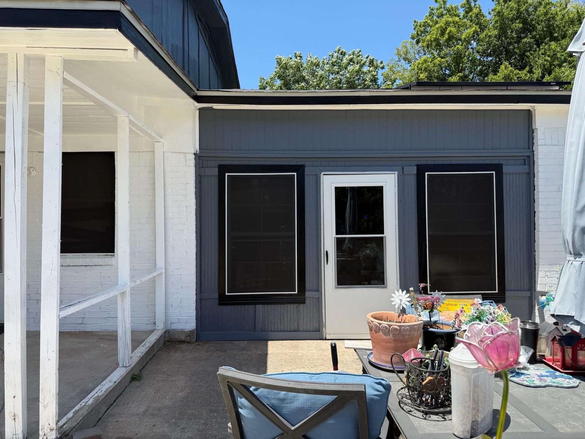 Back patio with white columns, dark-sided house wall, and a small table with potted plants and patio items.