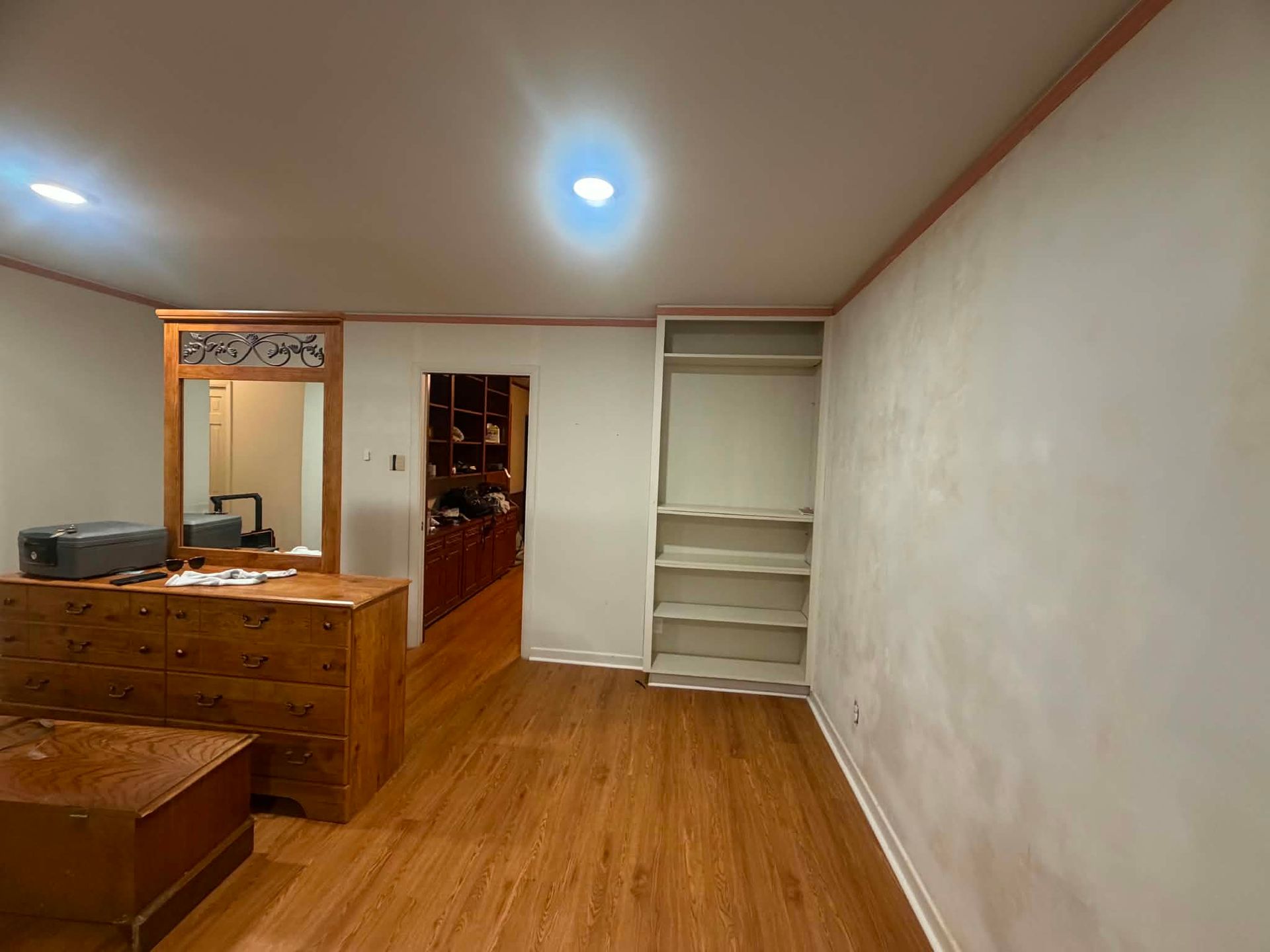 Empty bedroom with wood floors, dresser, mirror, doorway, and built-in shelves under ceiling lights