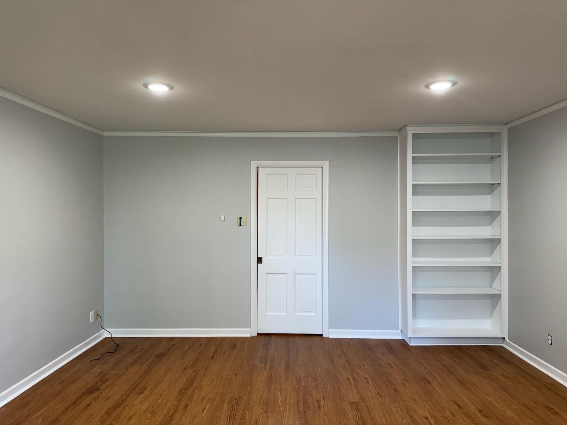 Empty room with gray walls, wood floor, white door, and built-in shelving