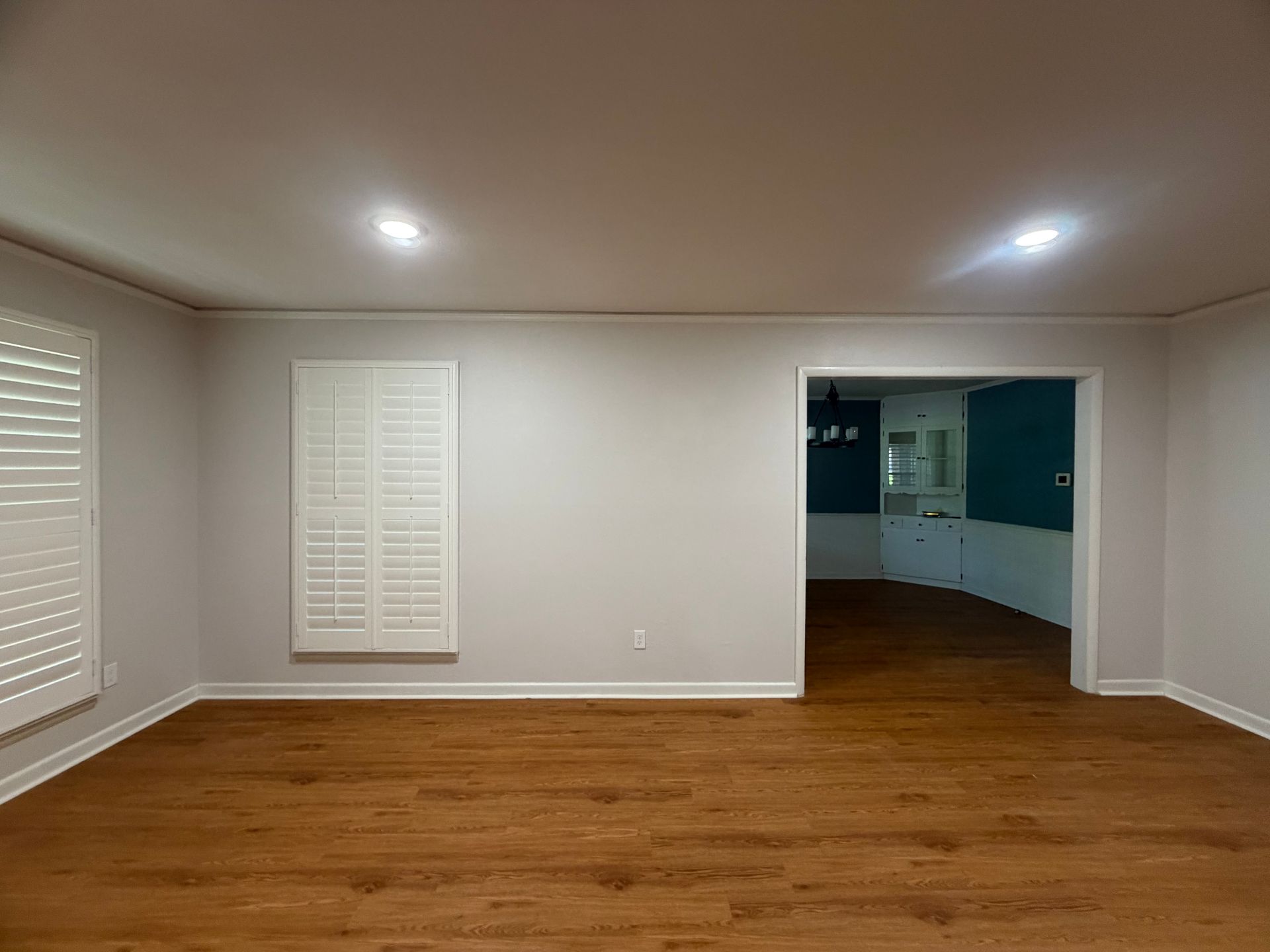 Empty living room with wood floor, white walls, and recessed ceiling lights, opening to a blue kitchen area.