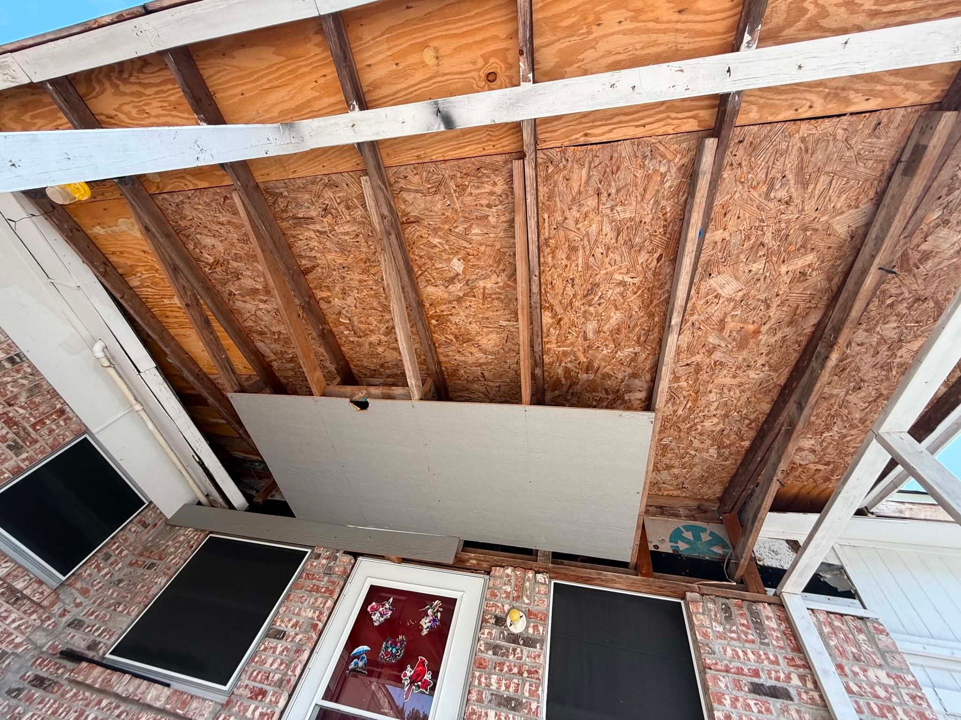 Partially finished ceiling with exposed wooden beams and a white panel over a red brick wall inside a room
