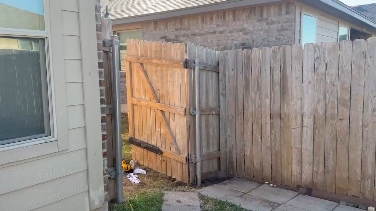 Wooden gate and fence beside a house, with a window and small plants along the concrete path.