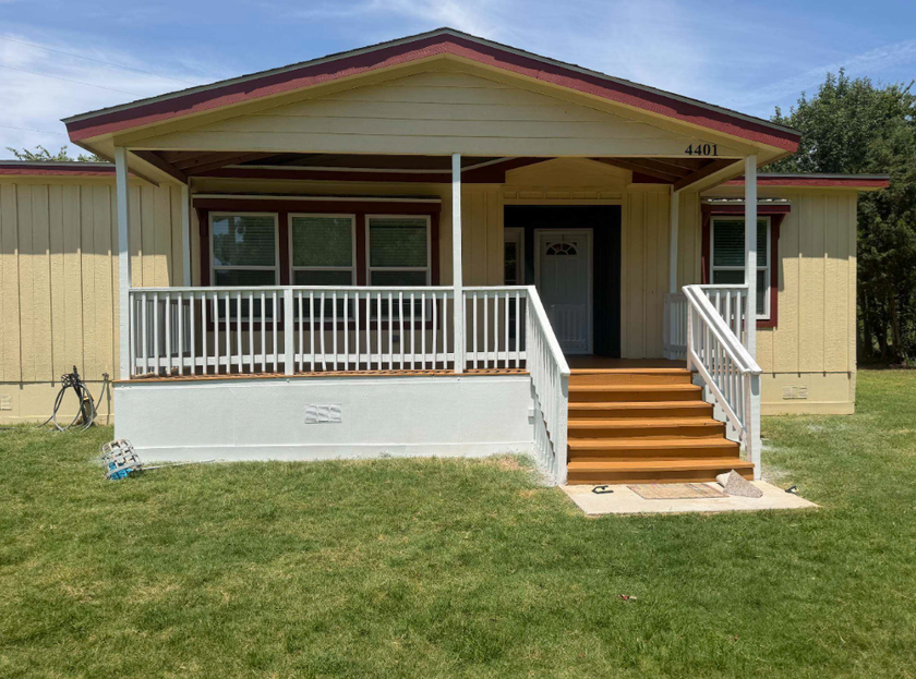 Small beige house with a front porch, white railings, and steps leading up from a grassy yard