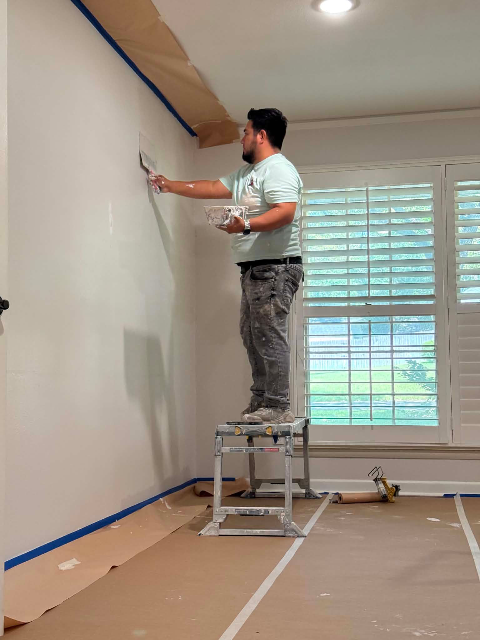 Person on a step stool painting a white wall indoors with blue painter’s tape nearby.