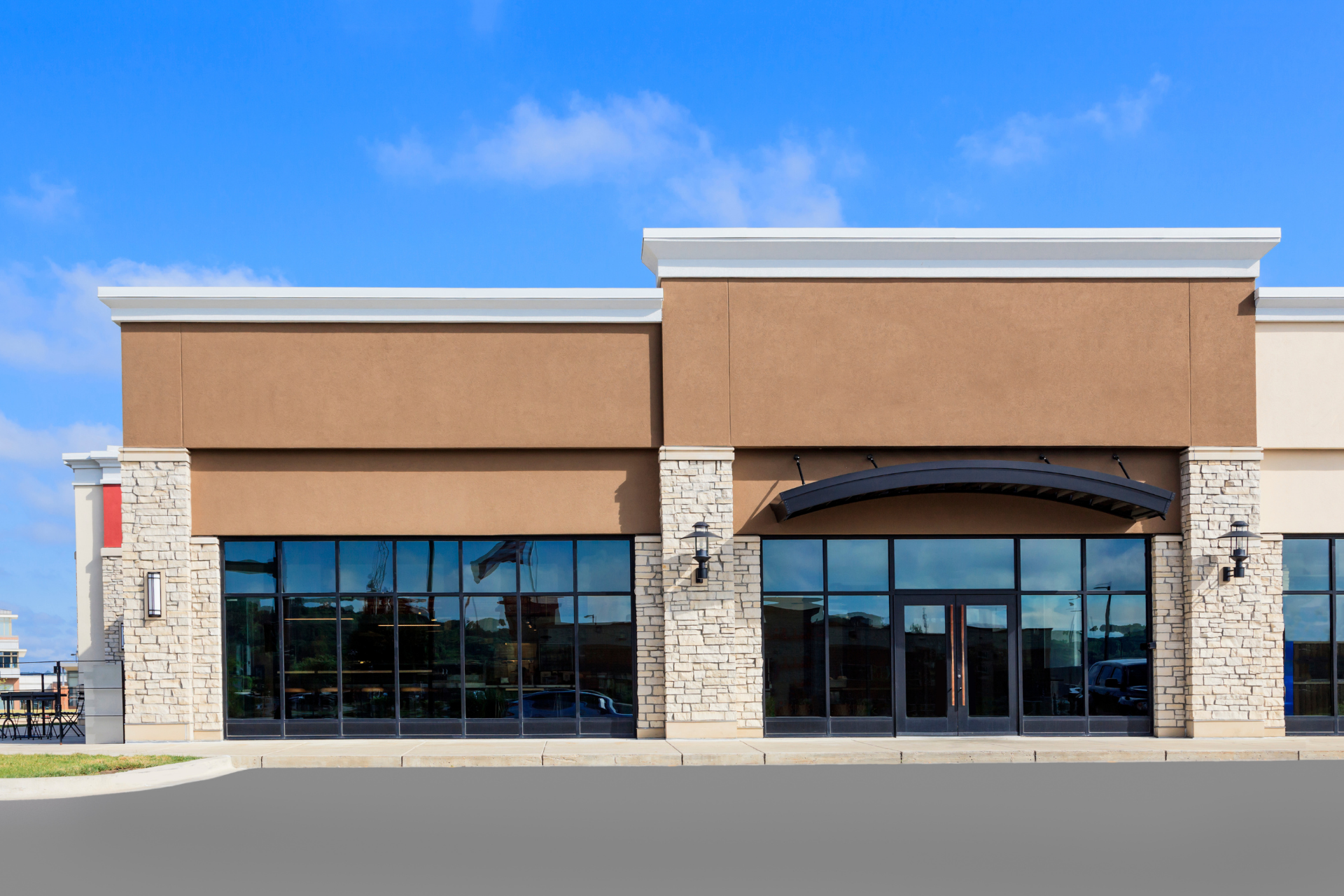 Empty retail storefront with tan facade, stone accents, and large glass windows under a blue sky