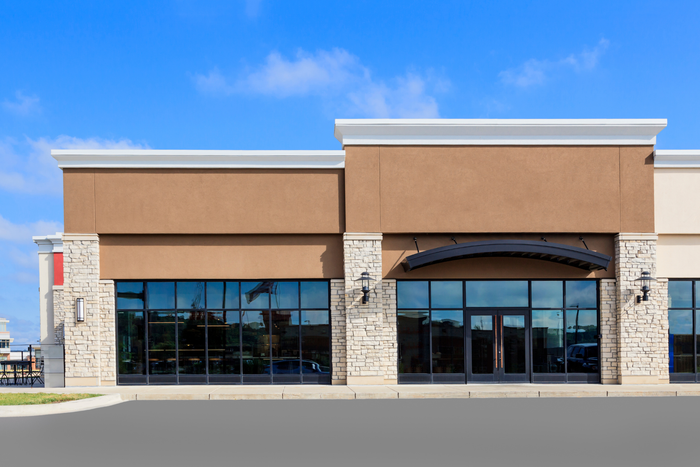 Empty storefront with tan facade, stone columns, and large glass windows under a blue sky