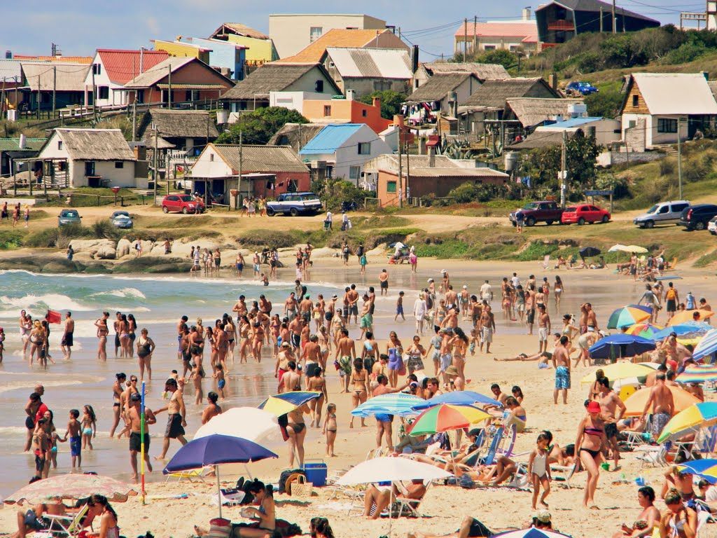 A beach filled with people and umbrellas with houses in the background