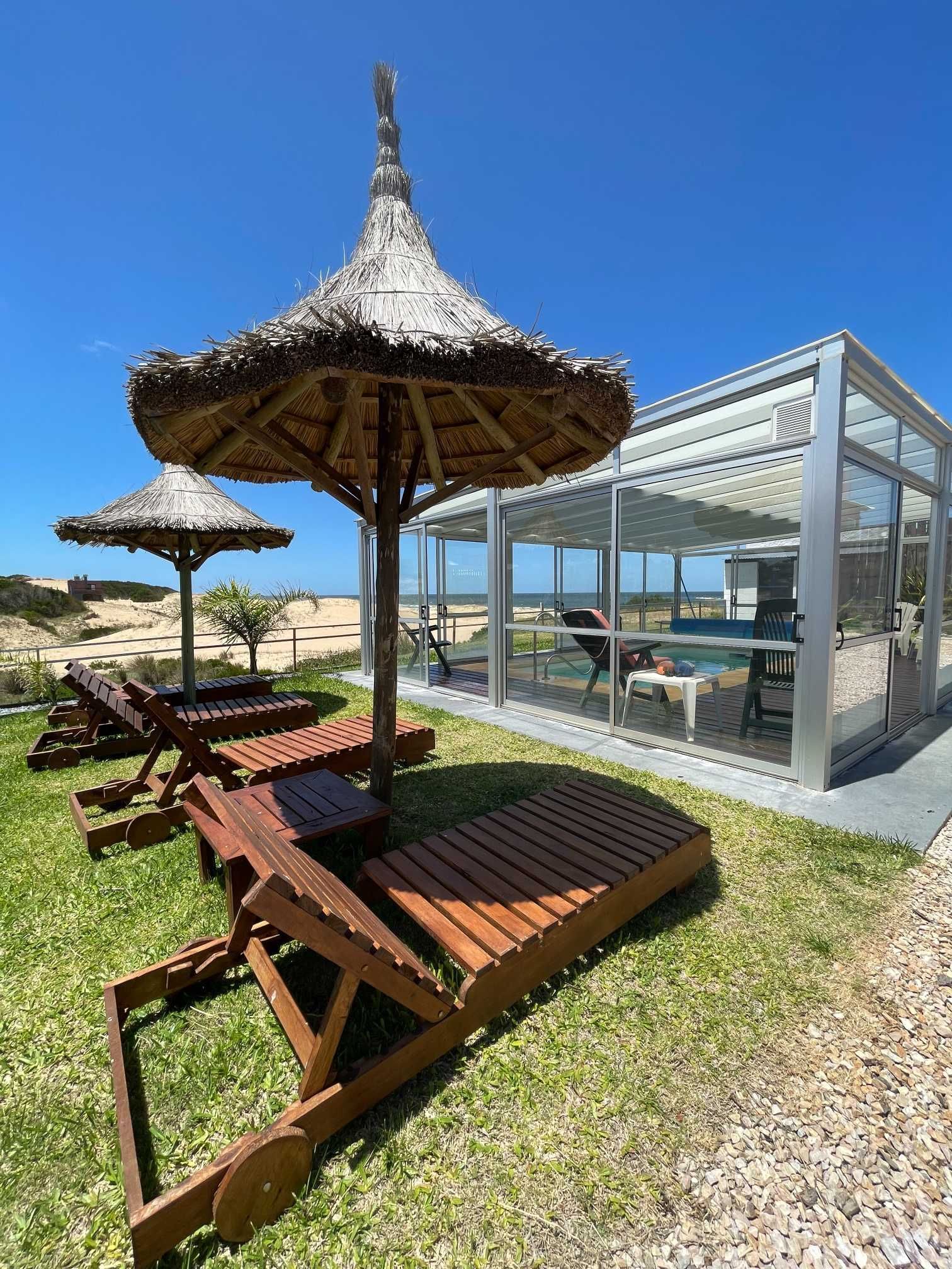 A row of wooden chairs and umbrellas in front of a swimming pool.