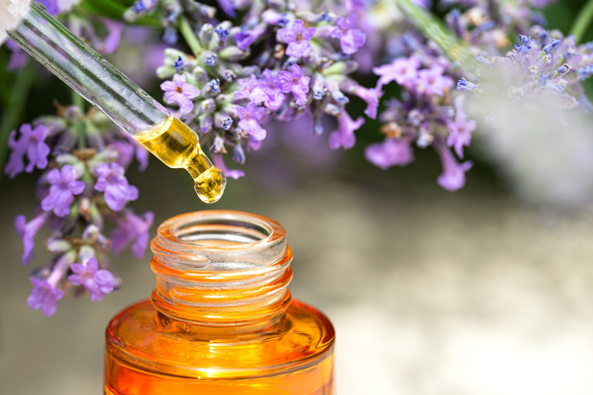 A drop of oil is being poured into a bottle with purple flowers in the background.