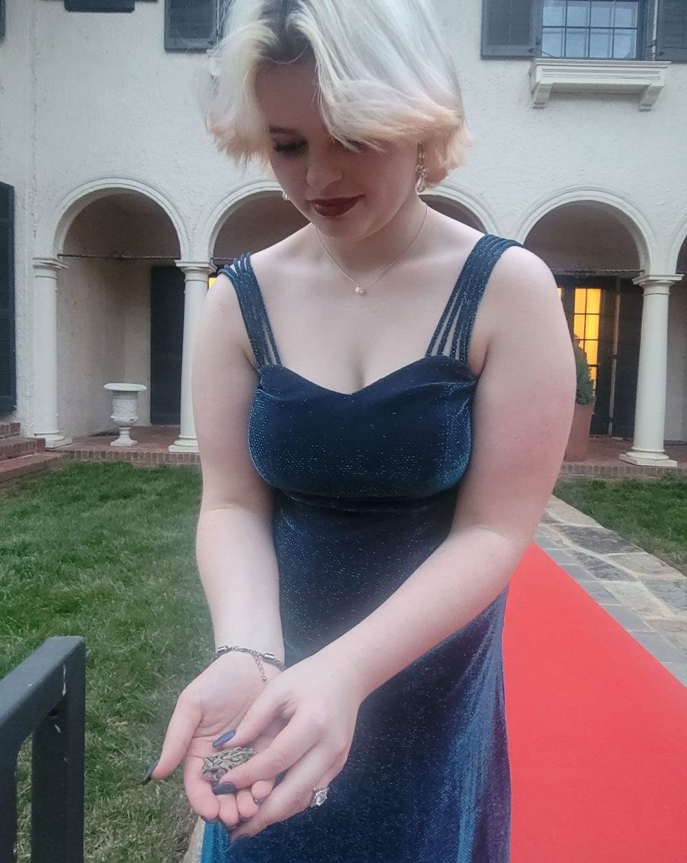 Person in a dark blue dress holding hands together on a red carpet in a courtyard.
