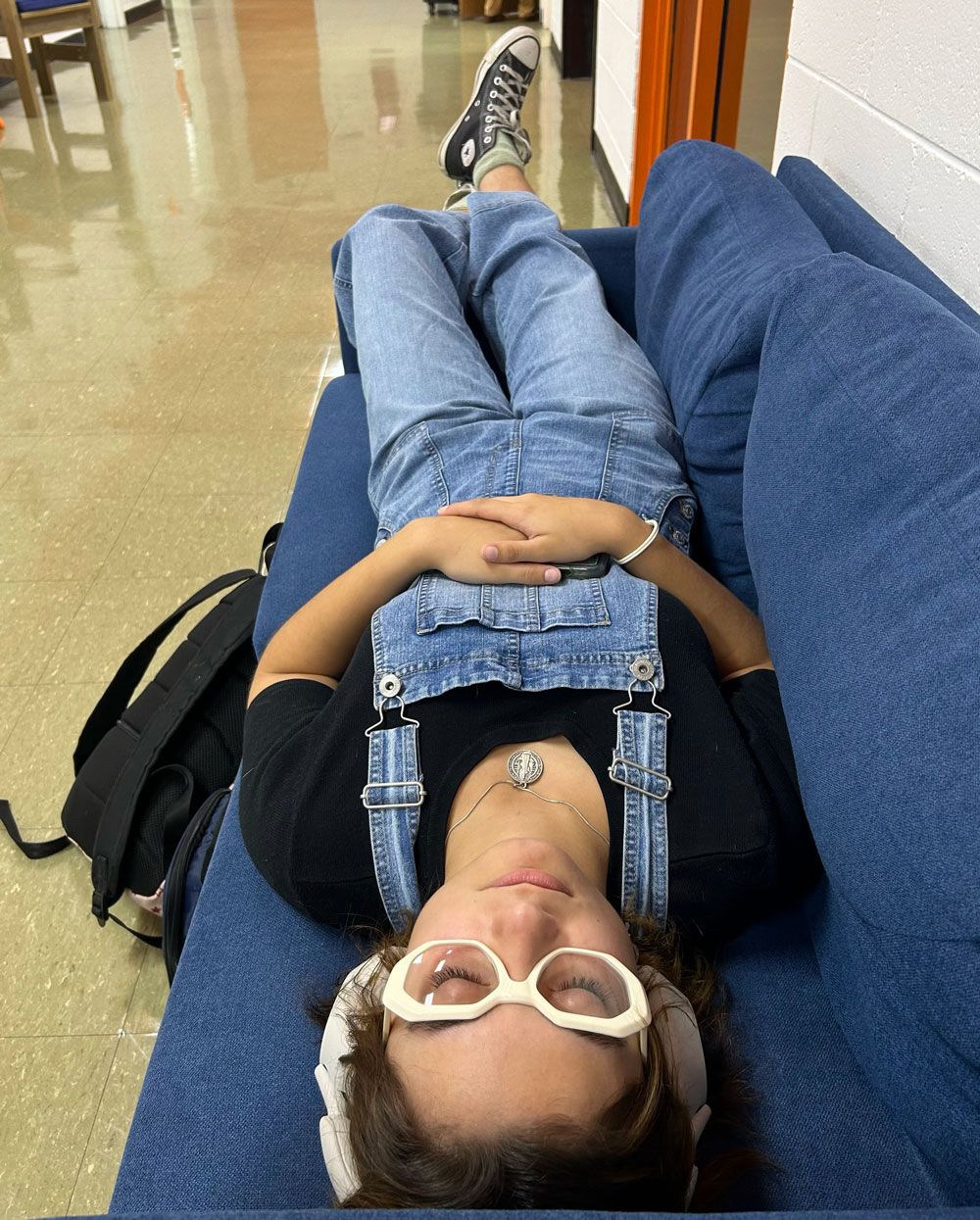 Person lying on a blue couch with white headphones, denim overalls, and a backpack beside them in a hallway.