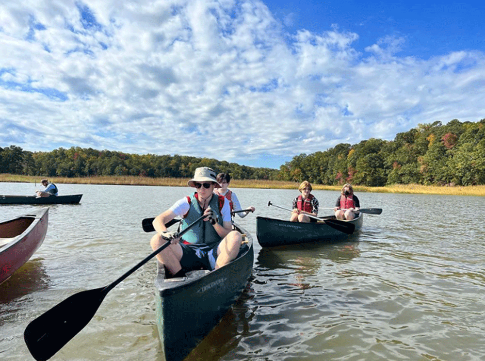 People canoeing on a calm lake, with blue sky, trees, and other boats in the background.
