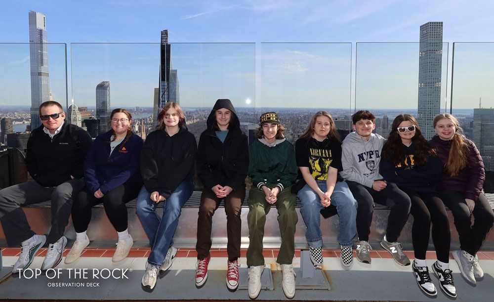 Eight people sitting on a ledge at an observation deck with a city skyline behind them.
