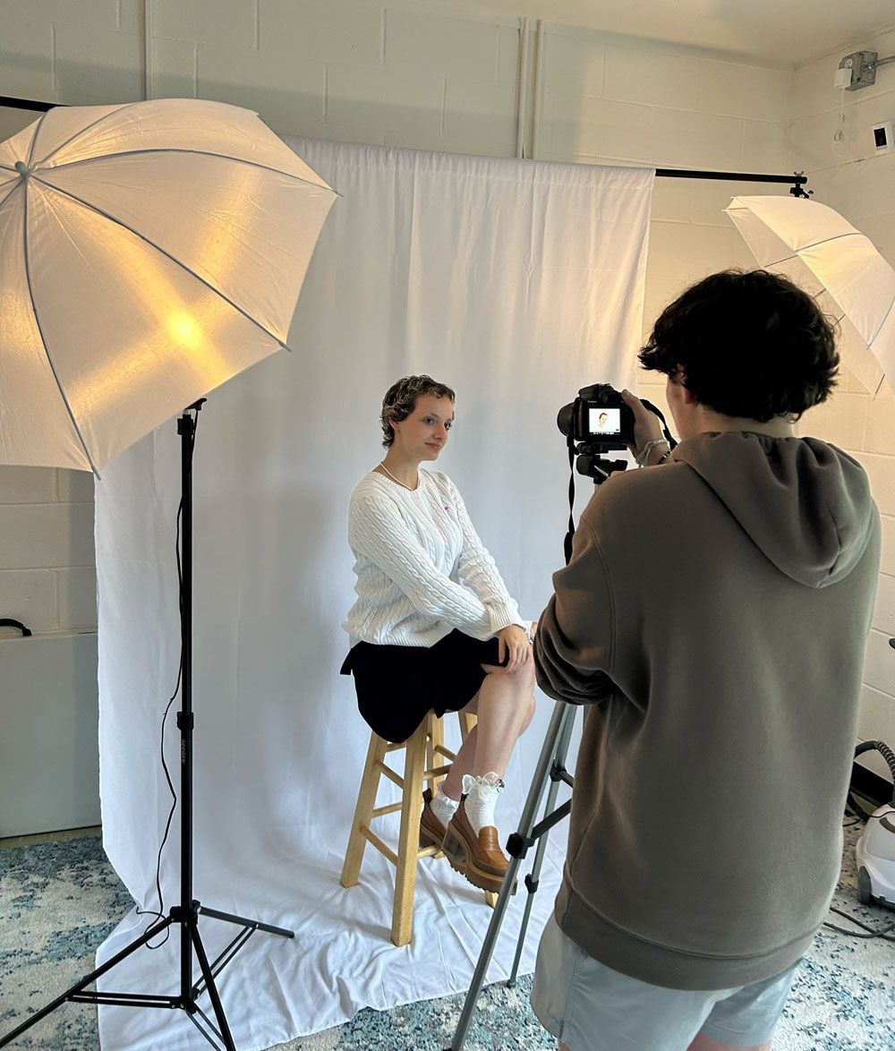 Photo shoot in a studio, with a seated model, photographer, and large lighting umbrellas against a white backdrop.