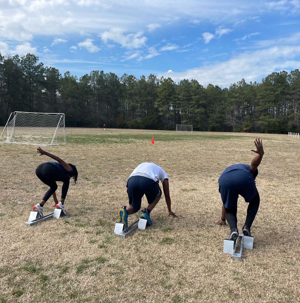 Three runners at starting blocks on a grassy field, about to sprint under a blue sky.