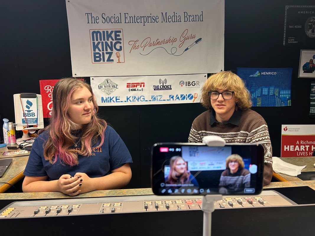 Two people sit at a table with a laptop in front of a Mike King Biz event banner.