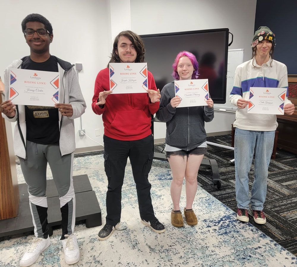 Four people holding certificates in a room, standing in front of a TV on a patterned rug.
