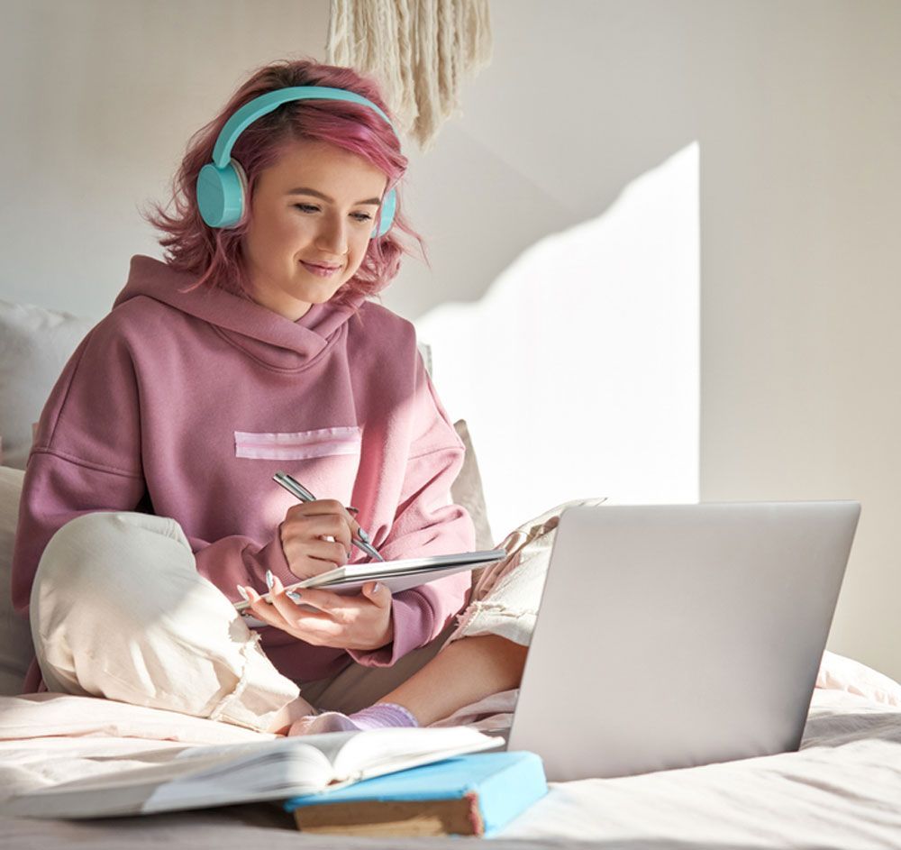 Image of a student on a bed working on a laptop with headphones on 