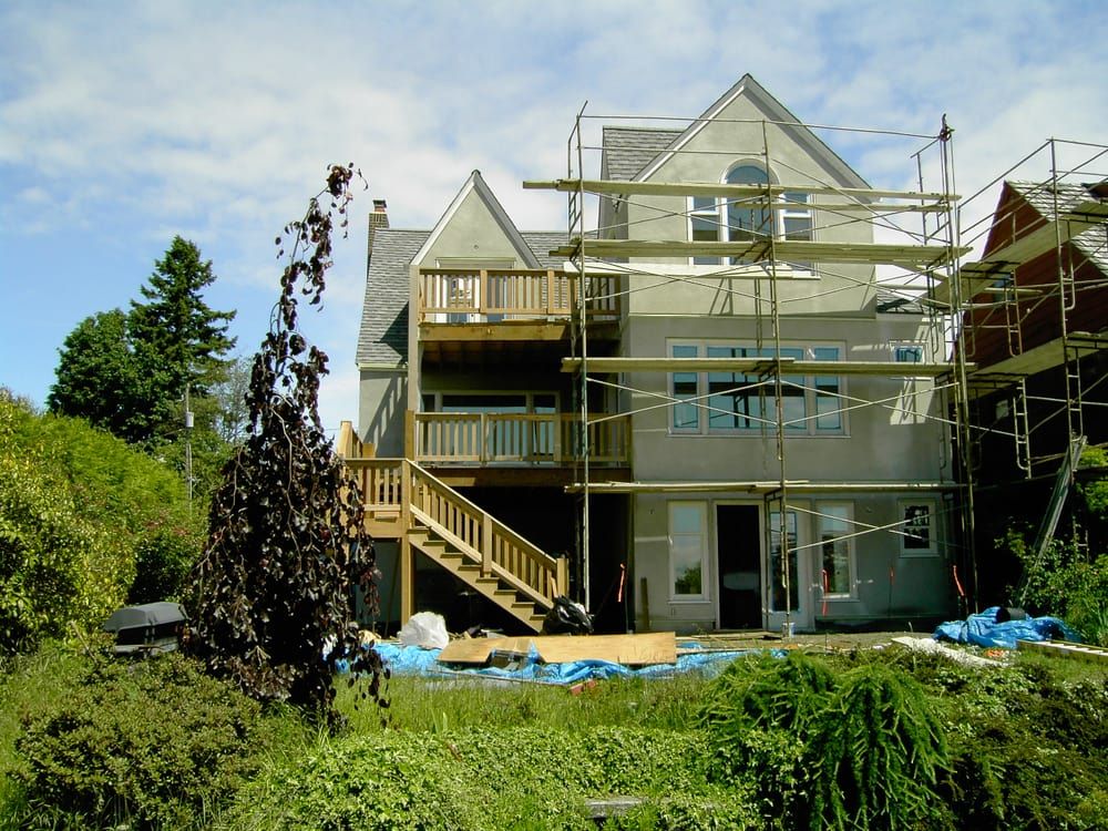 House exterior with scaffolding during renovation, wooden deck, stairs, blue tarps, green shrubbery.