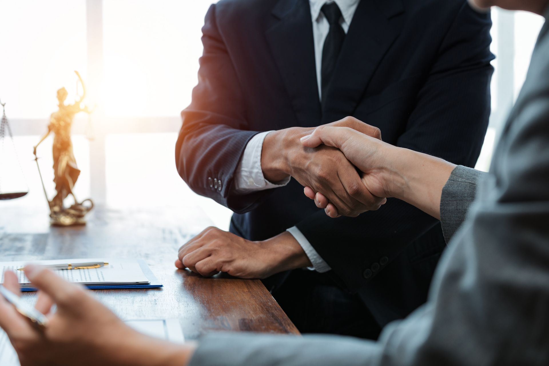 Businesspeople shaking hands across a table in a bright office, with a justice statue in the background