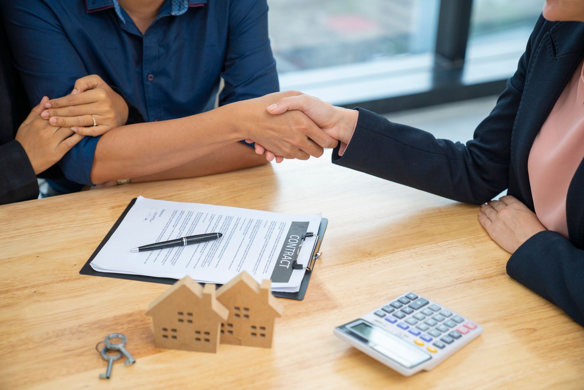 Two people shake hands over house documents with a calculator and house keys on a table