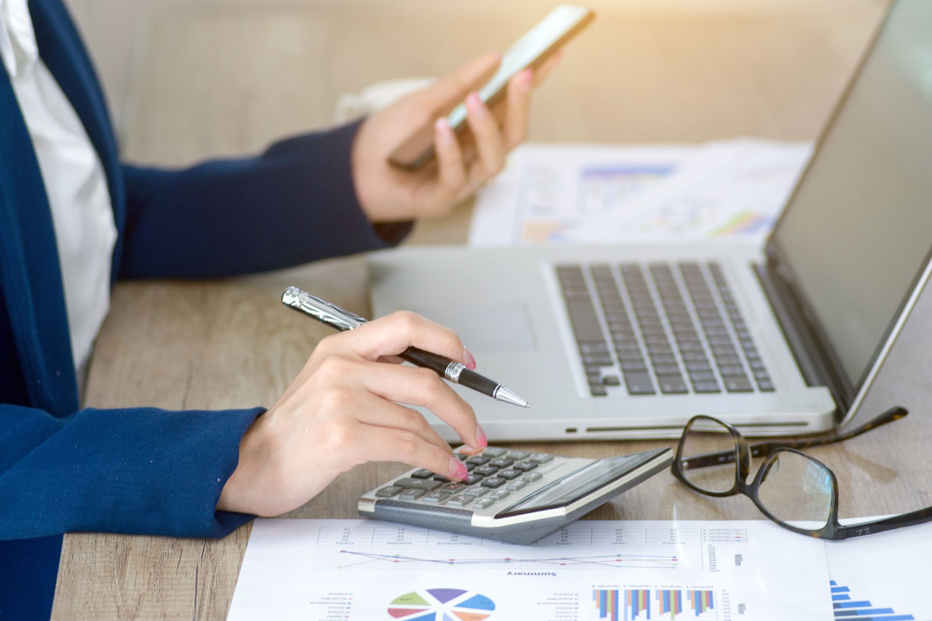 Hands using calculator and laptop at a desk with charts and eyeglasses