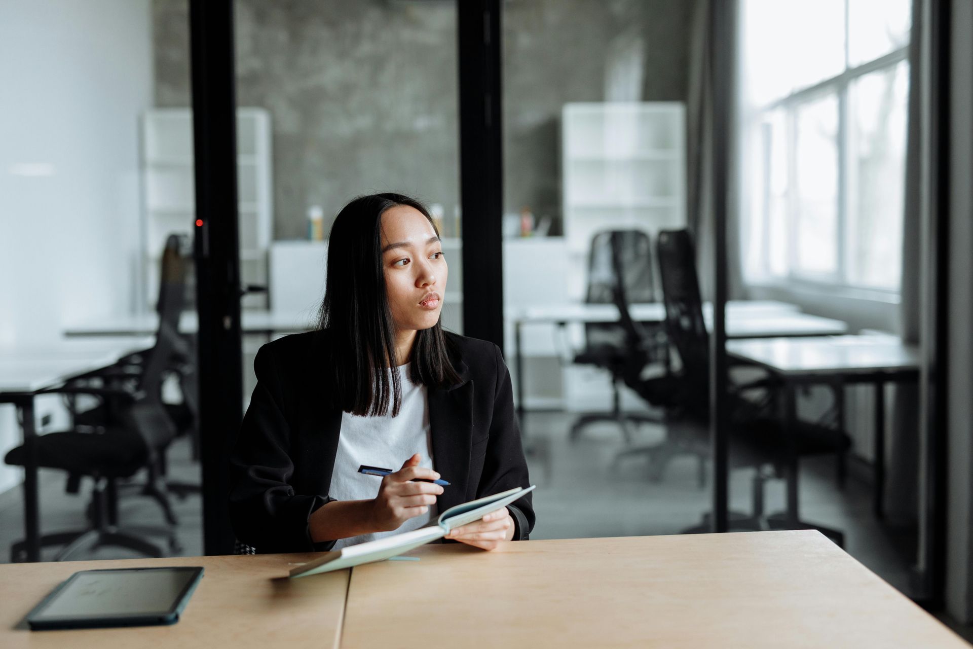 Woman in black blazer at desk, looking away, holding pen and tablet in office setting.