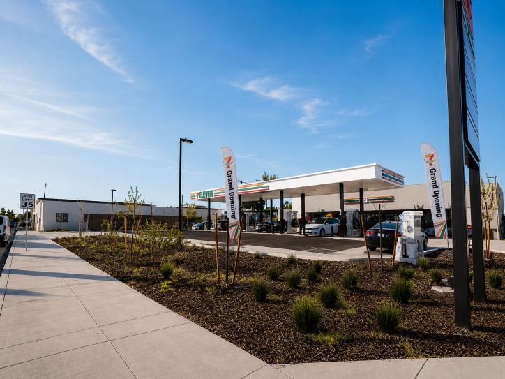 Gas station forecourt with pumps under a white canopy beside a sidewalk and landscaped planters under a blue sky
