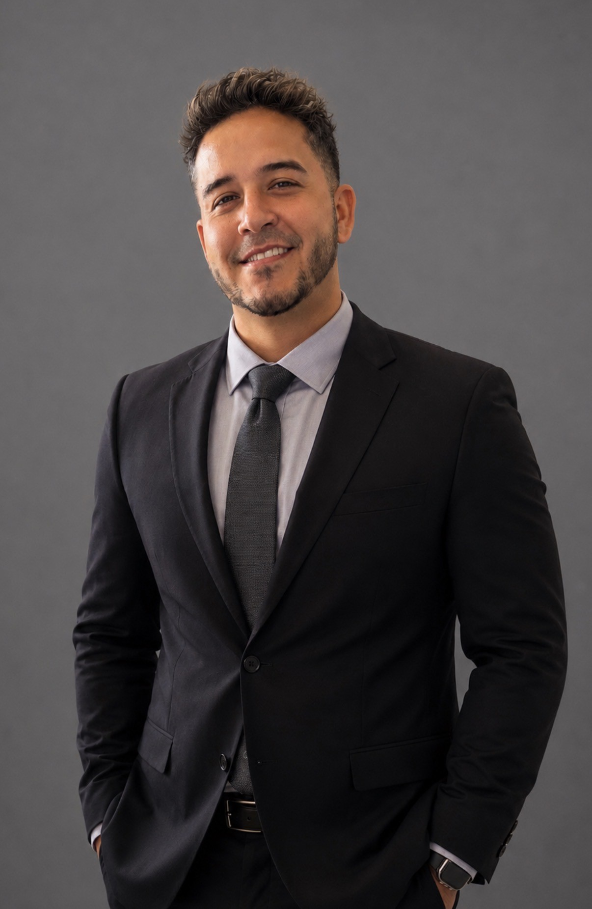 Smiling man in a black suit and tie against a gray background.