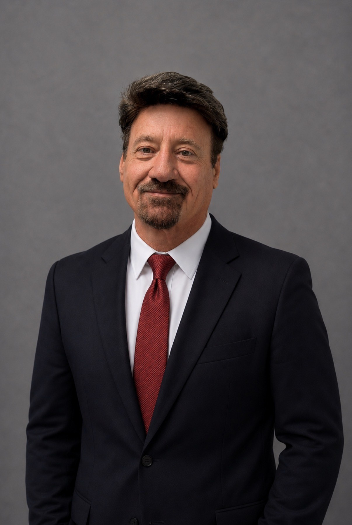 Man in a dark suit, white shirt, and red tie standing against a plain gray background.