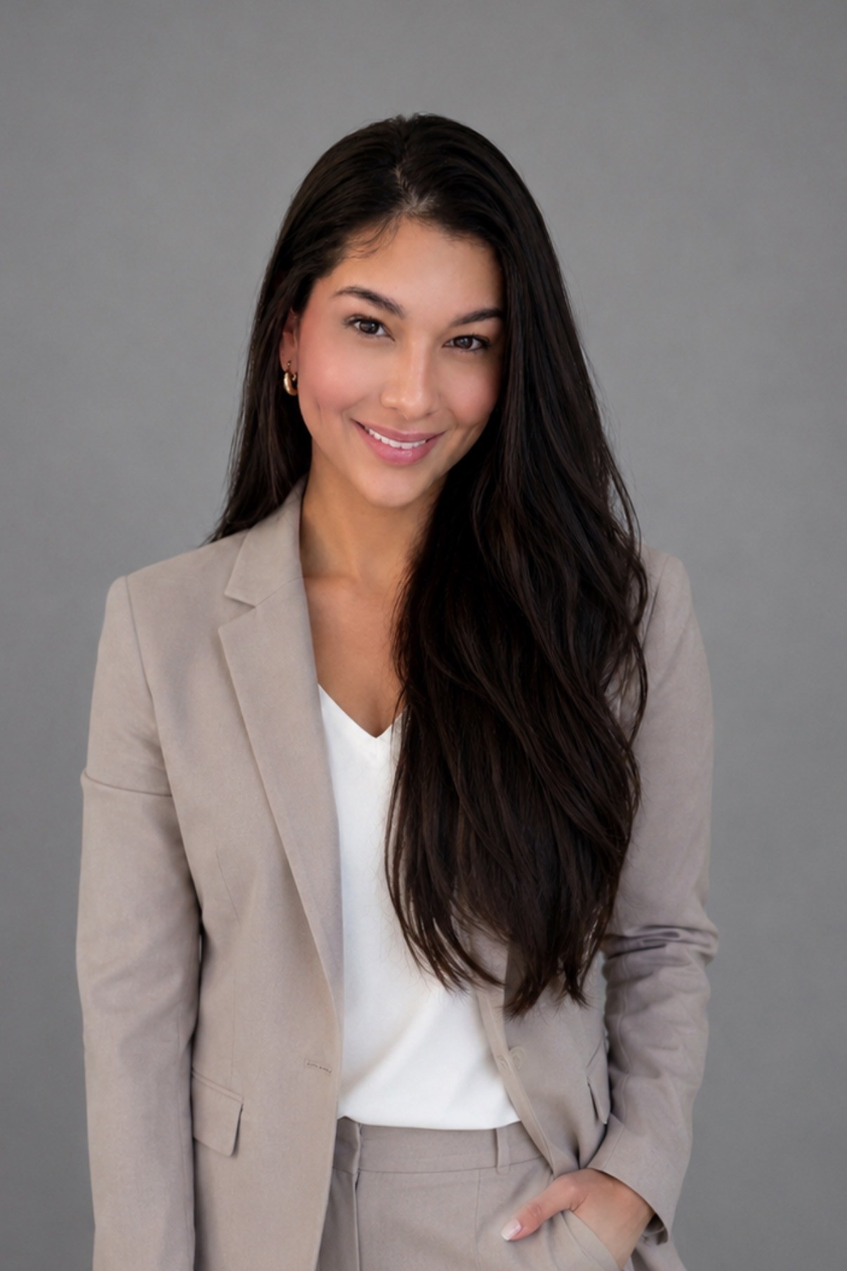 Woman in a beige blazer and white top, smiling against a gray background.