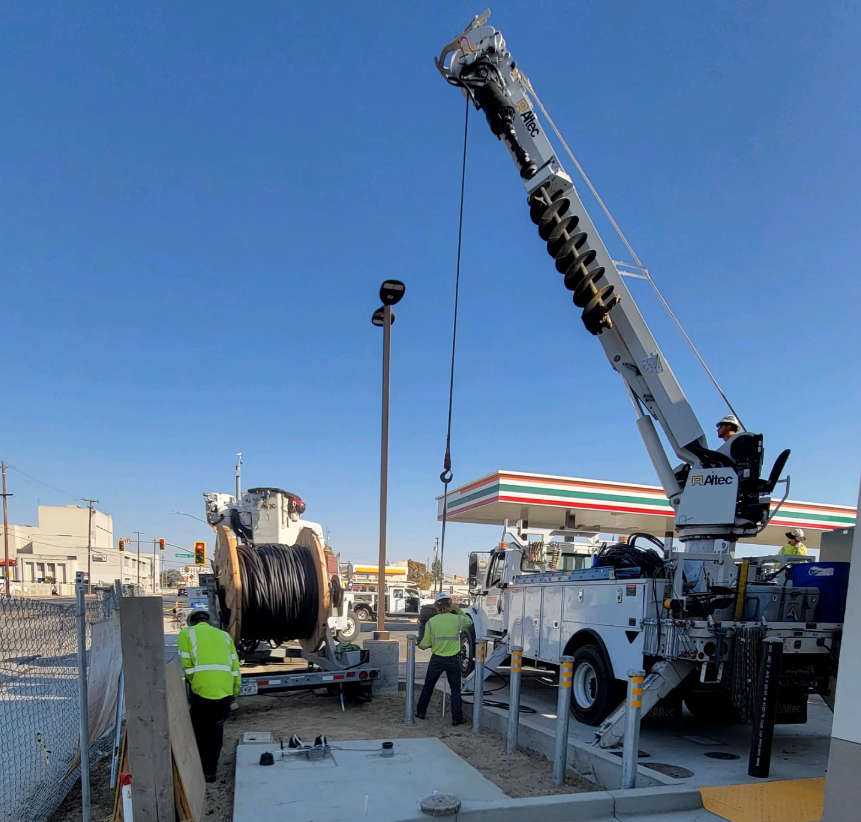 Crane truck lifting equipment near a 7-Eleven store; workers in yellow vests present.