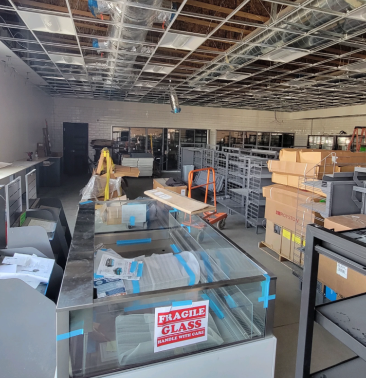 Interior of a commercial space under construction. Display cases, metal shelving, and boxes are visible. Open ceiling.