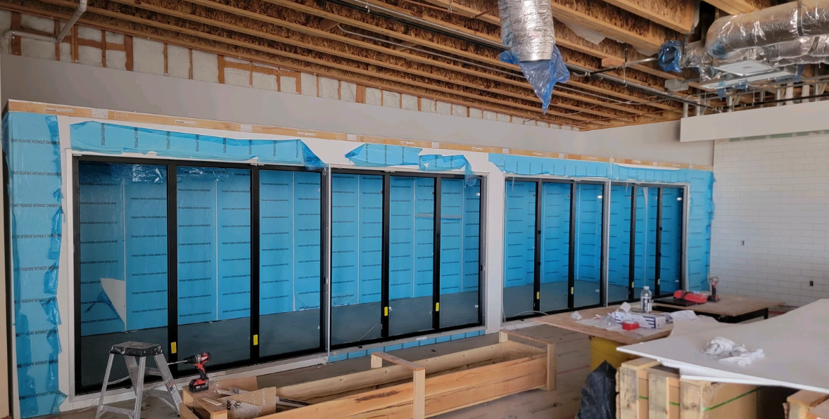 Construction site: large windows framed in black against blue insulation, unfinished ceiling and wood framing.