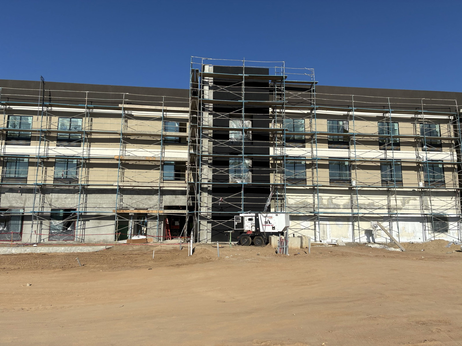 Scaffolding surrounds a multi-story building under construction on a dusty site under a clear sky
