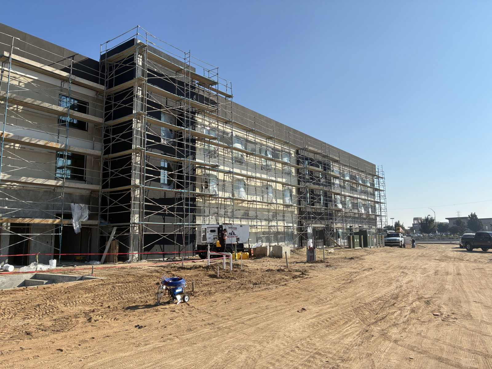 Scaffolded multi-story building under construction on a dirt lot under a clear blue sky