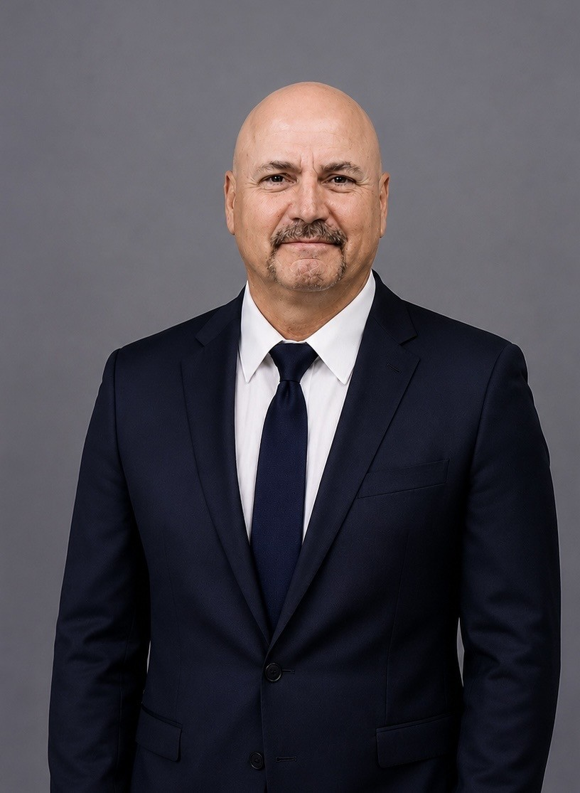 Man in a dark suit and tie standing against a gray studio backdrop, facing forward with a slight smile