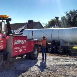 Red tow truck lifting a large, silver tank trailer. Man in orange shirt watches under a blue sky.