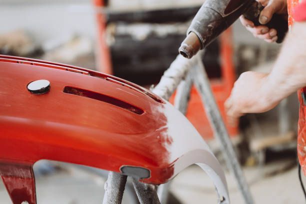 Close-up of a person working on a red car bumper, using a heat gun or similar tool for automotive repair or refinishing. The background shows workshop equipment, creating a busy repair shop environment.