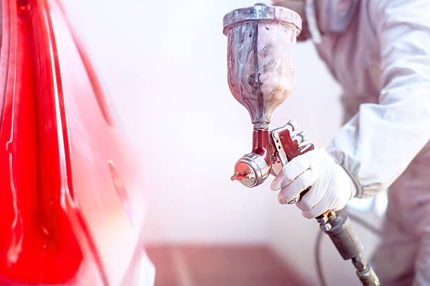 Person in protective gear spraying red paint onto a car surface with a spray gun.