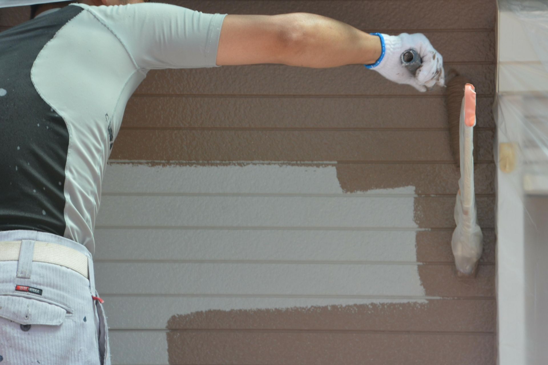Person painting siding on a house, wearing gloves, painting with a brush.