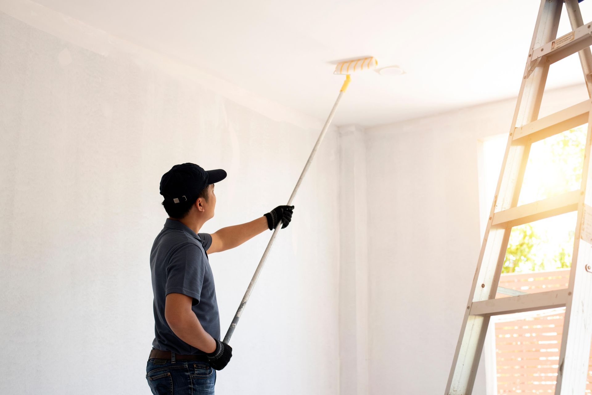 Person painting a ceiling with a roller on an extension pole, standing near a ladder in a bright room.