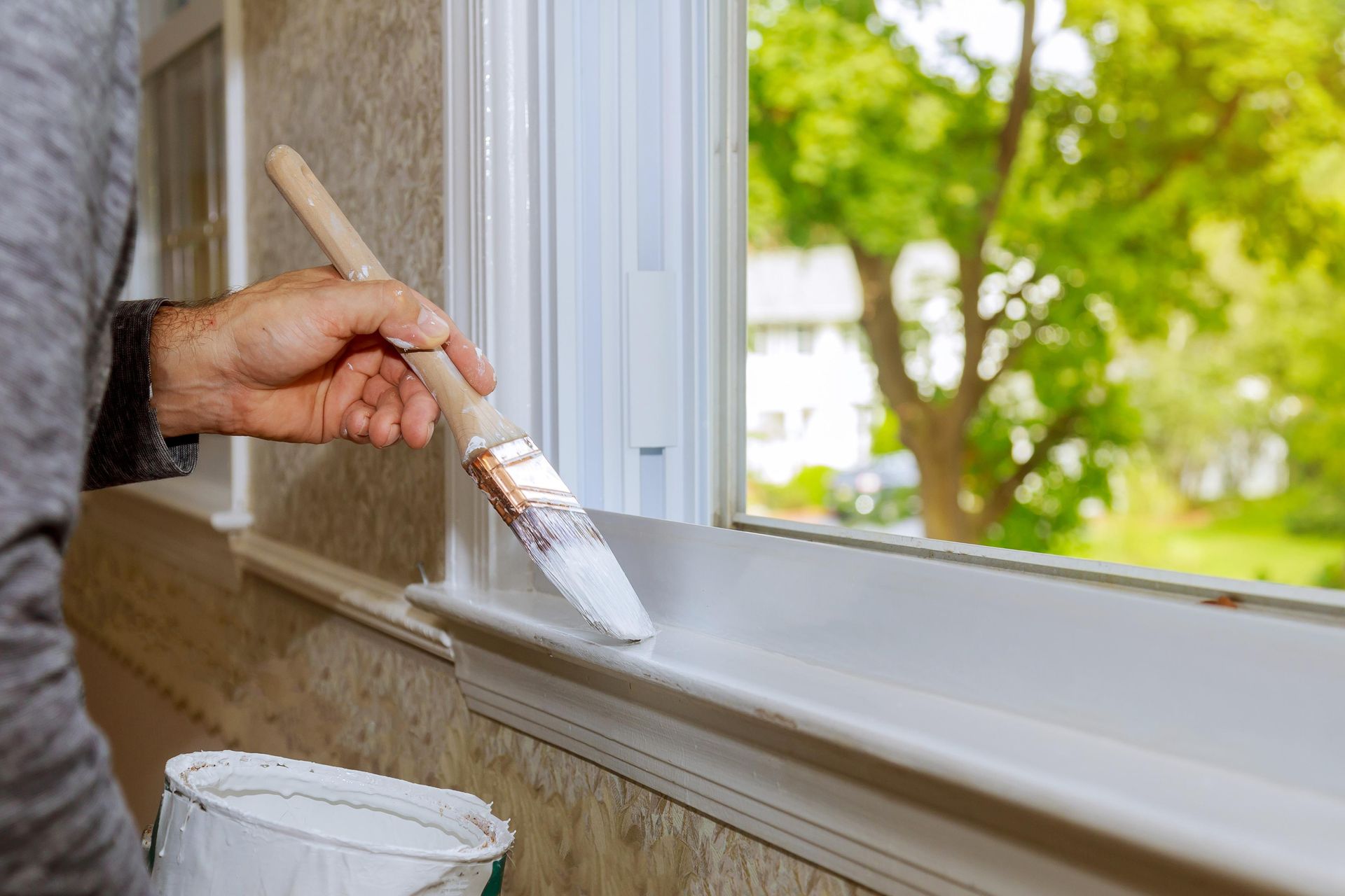Person painting a white windowsill with a brush; trees visible through the window.