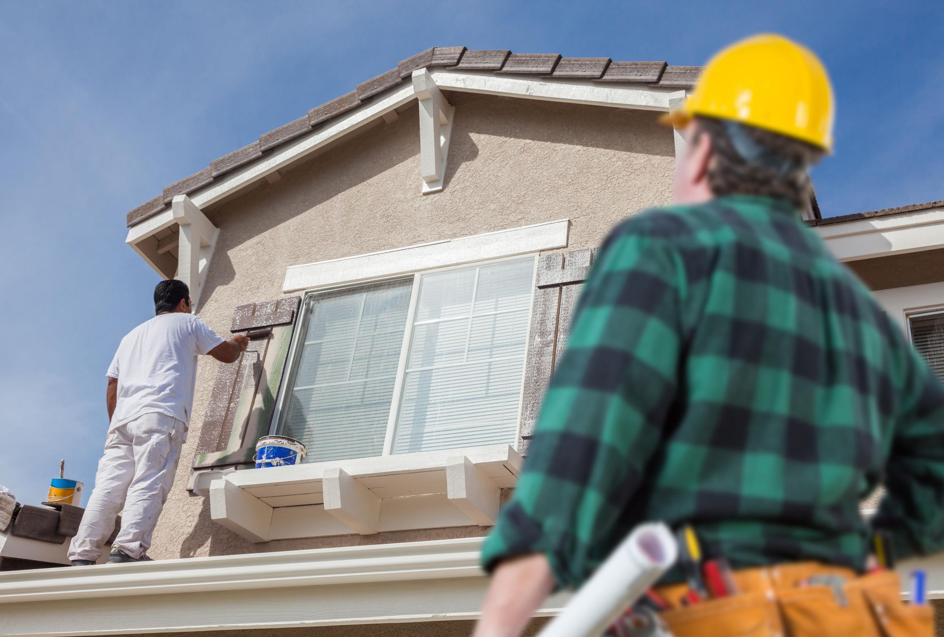 Two construction workers painting a house; one on roof, one in focus wearing yellow hard hat.